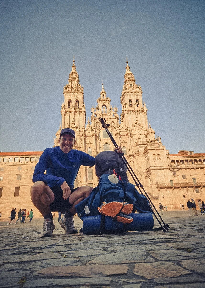 A smiling pilgrim crouches next to his fully loaded bike, which features the Camino symbol—a white scallop shell with a red cross—while standing in Obradoiro Square in front of the Santiago de Compostela Cathedral
