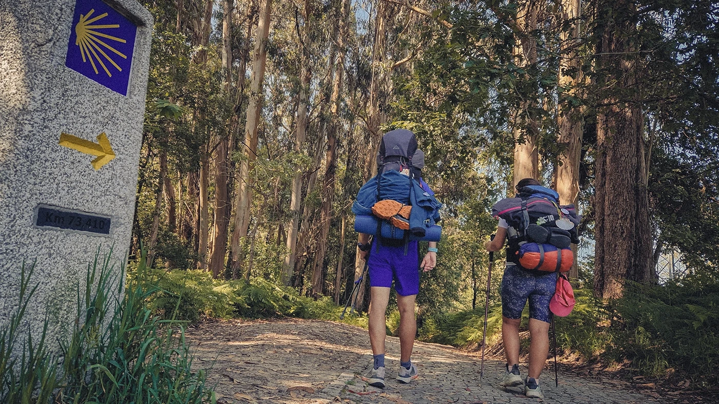 Two pilgrims with heavy backpacks walk through a forest in Galicia on their way to Santiago, guided by a white marble waymarker stone with an arrow pointing towards their destination