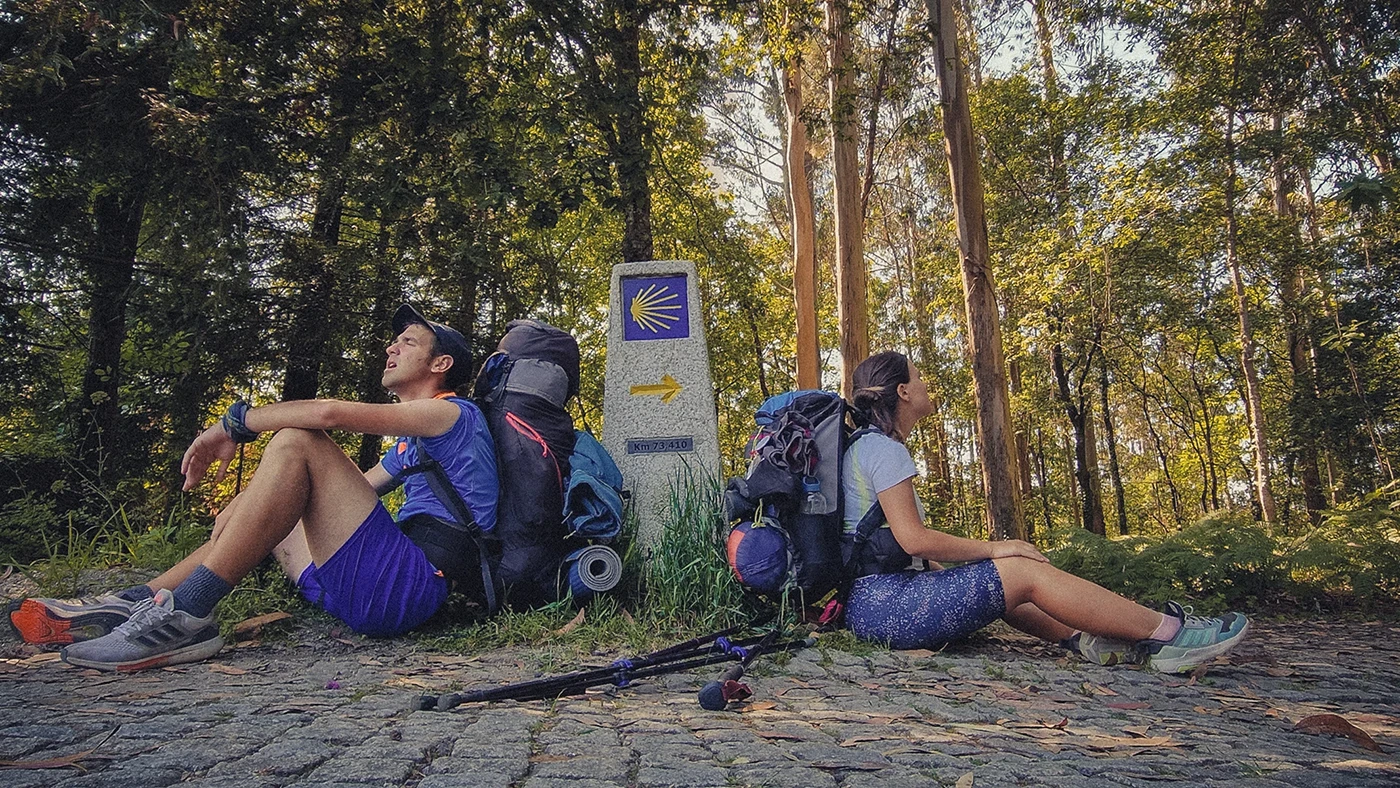 Two tired pilgrims rest against a waymarker stone in Galicia while hiking along the Camino de Santiago