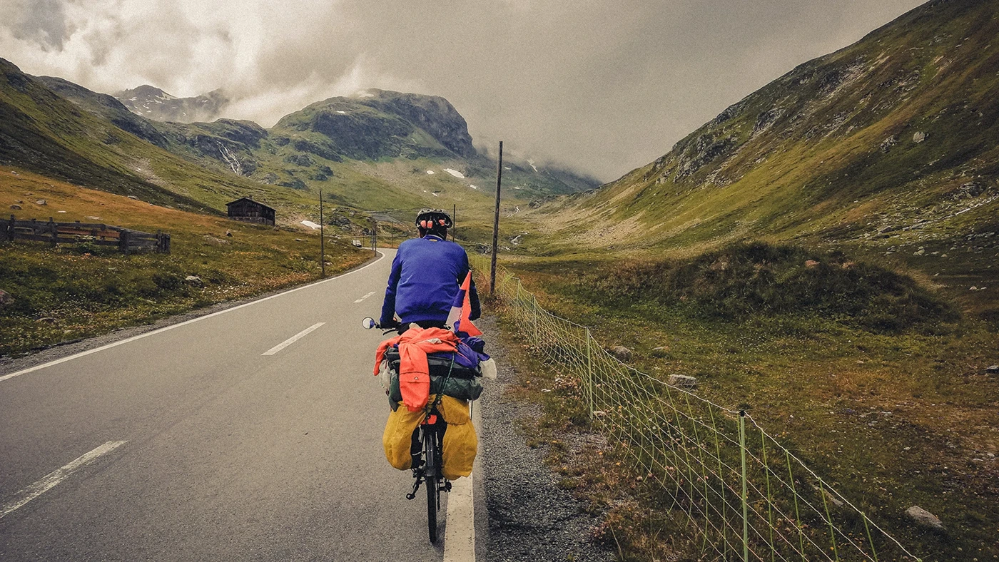 A rear-view shot of a fully loaded cyclist looking at the mountains and peaks of Flüela Pass in Switzerland, now enveloped in thick clouds.