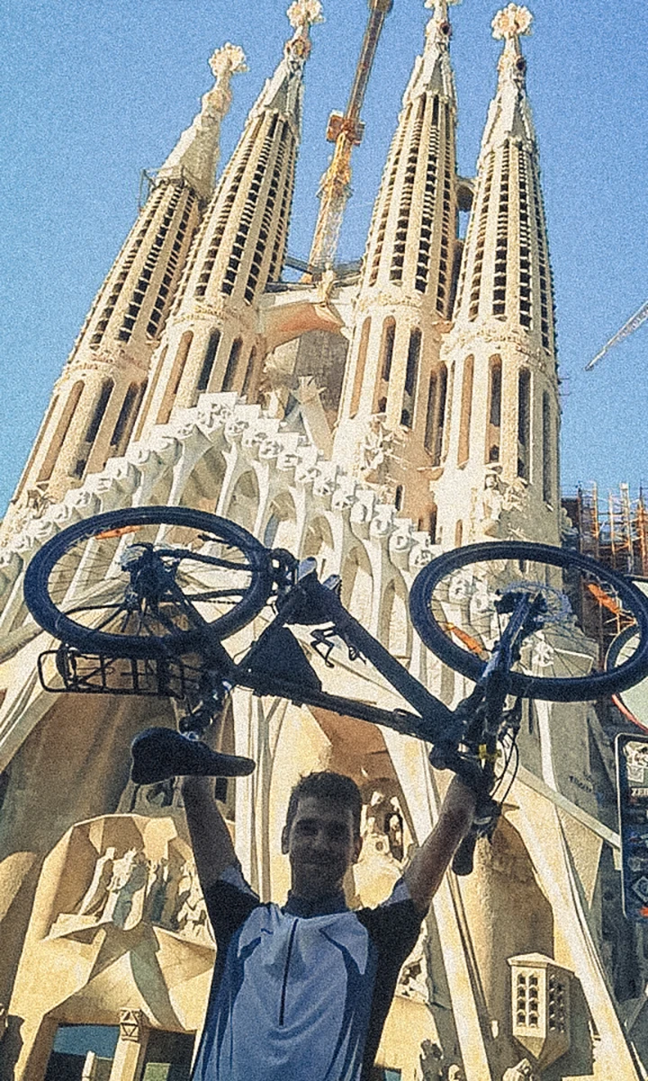 A smiling and satisfied cyclist triumphantly lifts his bike in front of Sagrada Familia in Barcelona, with the iconic basilica standing tall behind him