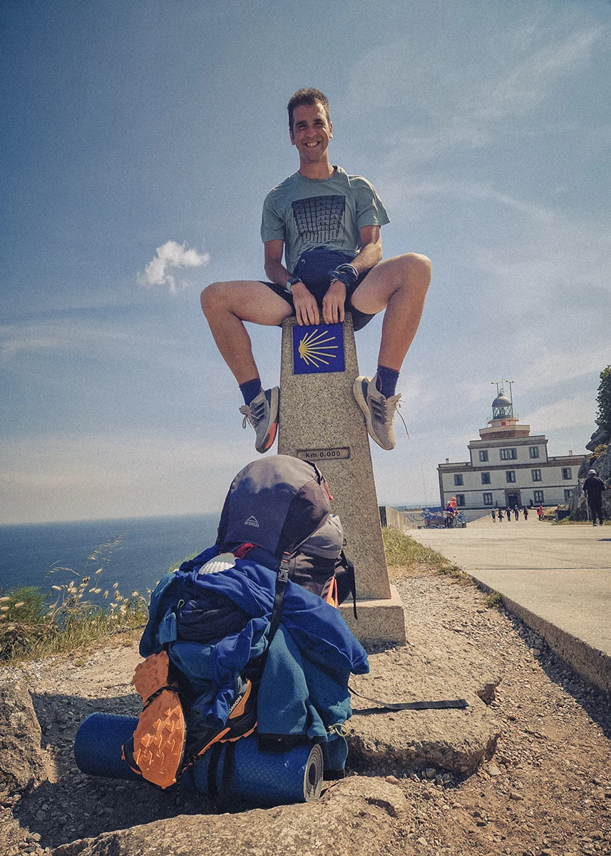 A smiling adventurer sits on the final milestone at Cape Finisterre, which marks 0.00 km, with his fully loaded backpack leaning against the stone. In the background, the rugged coastline and the Finisterre lighthouse stand against the horizon