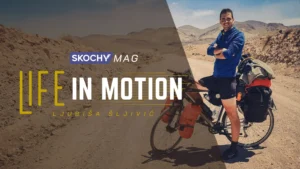 A smiling cyclist stands over his loaded bike, arms crossed, admiring the desert landscapes of Morocco.