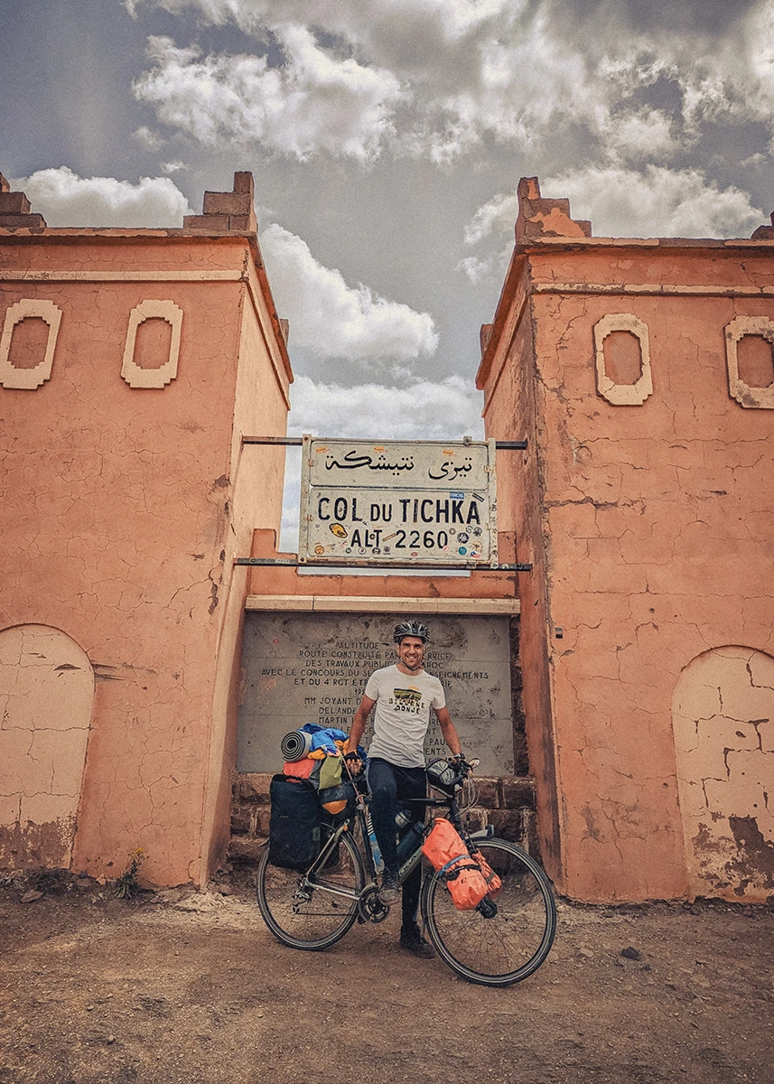 A smiling bikepacker stands with his fully loaded bicycle next to the monument at the Tizi n'Tichka pass in Morocco.