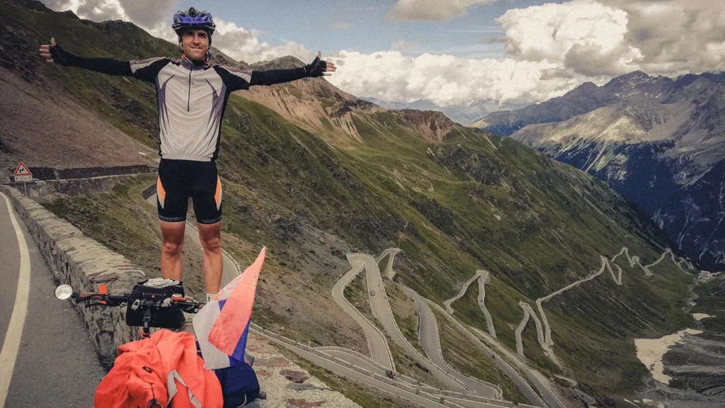 A joyful cyclist triumphantly spreads his arms beside his bike, celebrating the successful ascent of Passo dello Stelvio (2,758 m), the toughest climb in the Alps, with winding switchbacks visible below