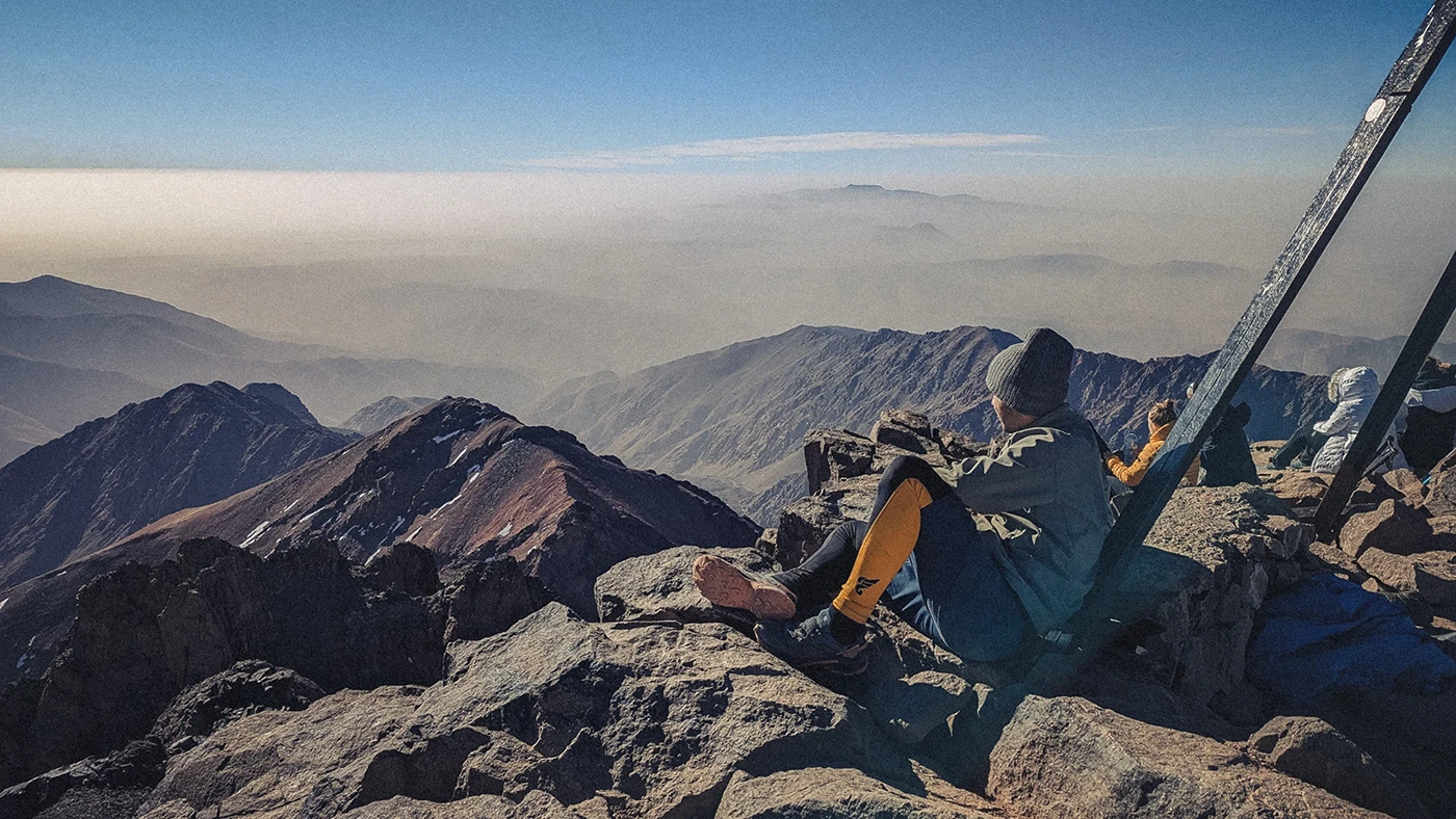A hiker sits atop Mount Toubkal, the highest peak in Morocco, leaning against a pyramid-shaped monument while admiring the breathtaking landscapes and surrounding peaks.