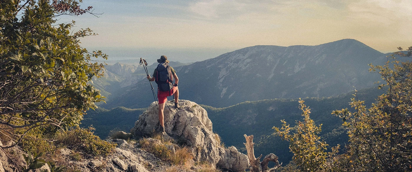 A hiker, leaning on his right knee with trekking poles in his left hand, admires the stunning Dalmatian landscape from a rocky summit.