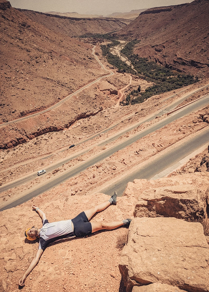 A fatigued cyclist lies on a rock, arms and legs spread out, overlooking the serpentines he just conquered in the Dades Canyon, High Atlas.